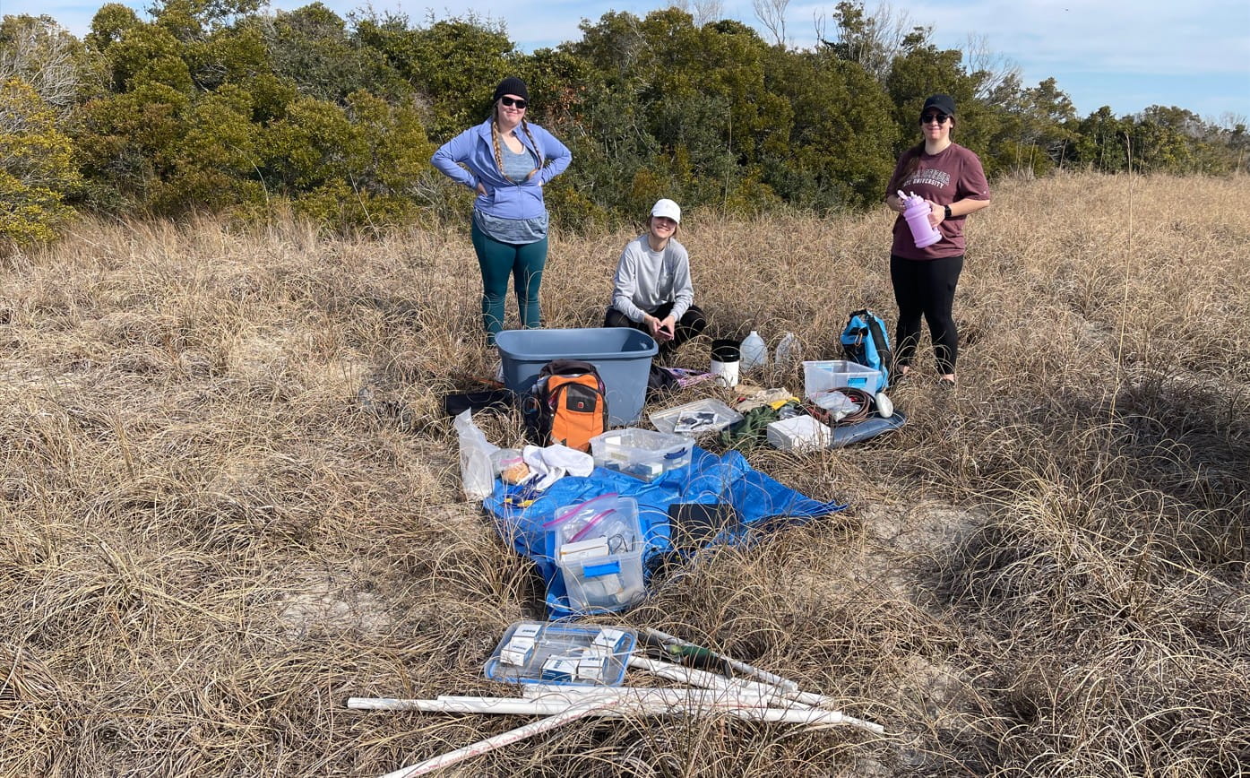 Three students outdoors next to a collection of field equipment unpacked on the grass