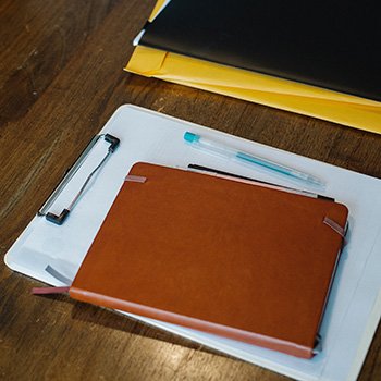 Photo of a journal on top of a clip board with a pen on top of it and a folder on top a stack of legal envelopes